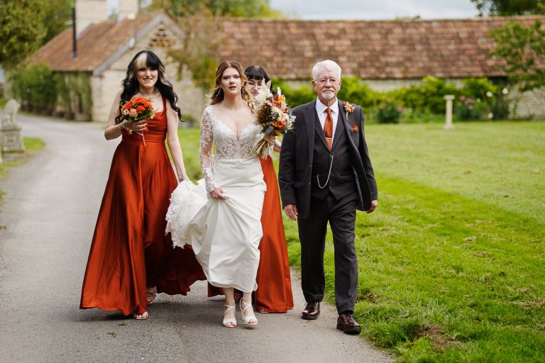Bride, bridesmaids in rust orange dresses, and father walking along stone cottage lane to outdoor ceremony at Pennard Hill Farm