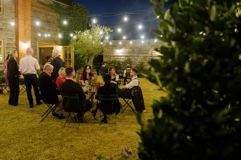 Intimate wedding party relaxing under warm festoon lighting in the stone courtyard at Pennard Hill Farm at dusk